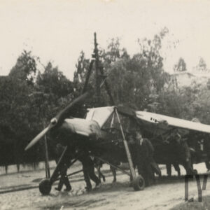 grupa mężczyzn pchająca samolot brukowaną ulicą, w głębi drzewa i zarysy budynków mieszkalnych