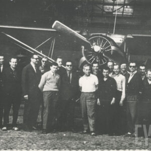 grupa mężczyzn, część ubrana w stroje robocze, część w garniturach, pozująca do fotografii przed hangarem z samolotem