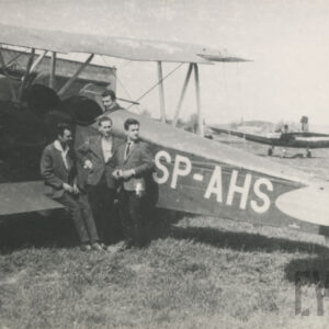 grupa mężczyzn w garniturach pozująca do fotografii przy dwupłatowcu stojącym na trawiastym lądowisku