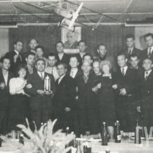 grupa elegancko ubranych osób pozujących przy zastawionym stole, nad głowami zgromadzonych wisi model samolotu