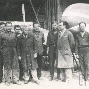 grupa mężczyzn w strojach roboczych, jeden z nich stoi ubrany w garnitur, pozujący do fotografii przy samolocie