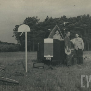 trójka mężczyzn stojąca przy pojeździe mechanicznym ustawionym na trawiastym lądowisku, w tle drzewa