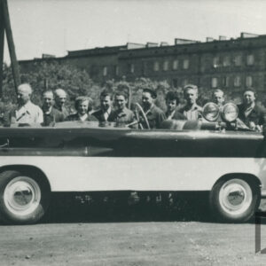 grupa osób w strojach roboczych pozująca do fotografii przy pojeździe mechanicznym na tle krzaków i budynków z cegieł