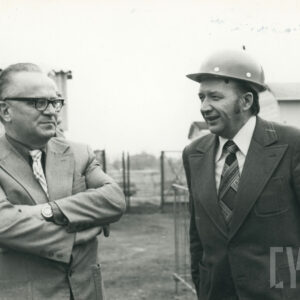 dwóch elegancko ubranych mężczyzn w garniturach, jeden z nich z kaskiem budowlanym na głowie, na tle budynków przemysłowych