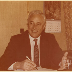 starszy mężczyzna w garniturze, siedzący przy biurku z piórem w dłoni