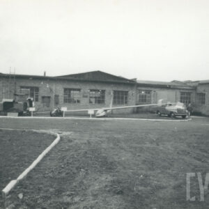 szybowiec i dwa pojazdy mechaniczne stojące na ziemi, otoczone budynkami przemysłowymi z cegły