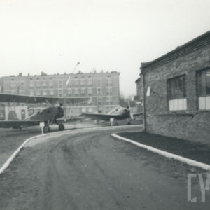 samoloty stojące na wolnej przestrzeni, w tle budynki mieszkalne z cegły