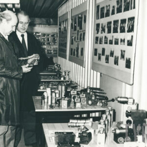 dwóch mężczyzn w garniturach pochylających się nad stołem z elementami metalowymi