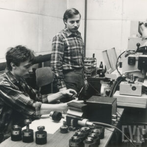 dwóch mężczyzn w strojach roboczych, jeden siedzi, drugi stoi przy biurku, na którym stoi urządzenie mechaniczne