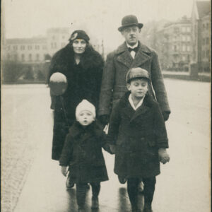 mężczyzna i kobieta w średnim wieku z dwójką młodych chłopców, wszyscy ubrani w eleganckie płaszcze i nakrycia głowy, spacerująca po chodniku, w tle kamienice i duży budynek z cegły