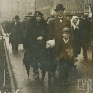 mężczyzna i kobieta w średnim wieku z dwójką młodych chłopców, wszyscy ubrani w eleganckie płaszcze i nakrycia głowy, spacerująca po chodniku, w tle kamienice i duży budynek z cegły