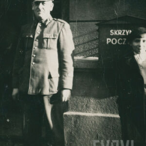 starszy mężczyzna w mundurze pocztowym, stojący obok skrzynki pocztowej na tle budynku z kamienia