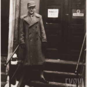 młody mężczyzna w płaszczu i czapce na głowie stojący na schodach przed dużymi, drewnianymi drzwiami, na drzwiach widoczna swastyka 