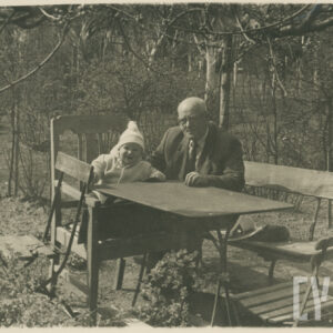 mężczyzna w starszym wieku ubrany w marynarkę, koszulę i krawat siedzący przy stole obok młodego chłopca w kurtce i czapce, w tle bezlistne drzewa