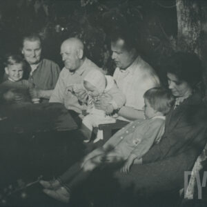 grupa osób: mężczyzna i kobieta w starszym wieku, mężczyzna i kobieta w średnim wieku oraz trójka małych dzieci, wszyscy siedzą przy stole w ogrodzie