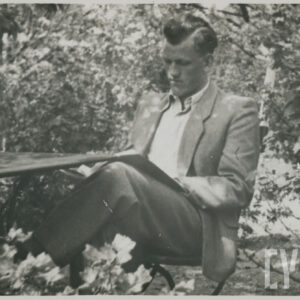 młody mężczyzna ubrany w koszulę i marynarkę, siedzący przy stoliku w ogrodzie, czytający książkę