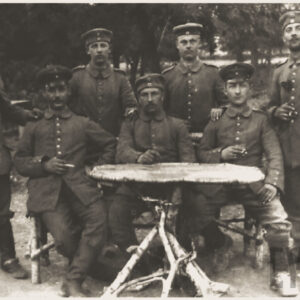 grupa młodych mężczyzn w mundurach wojskowych i czapkach na głowach, część z nich siedzi przy drewnianym stole, pozostali stoją