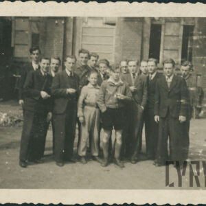 grupa młodych mężczyzn ubranych w większości w marynarki, koszule i krawaty, w głębi zniszczone budynki z cegieł