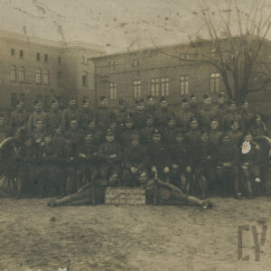 duża grupa młodych mężczyzn w mundurach i czapkach wojskowych pozująca do pamiątkowej fotografii na tle dużych ceglanych budynków, po bokach widoczne działa artyleryjskie