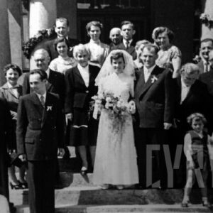 grupa elegancko ubranych ludzi otaczająca mężczyznę w garniturze i kobietę w sukni ślubnej na tle filarów budynku