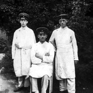 młodzi mężczyźni w ubraniach szpitalnych i czapkach wojskowych na głowach pozujący do fotografii, dwóch z nich stoi, a trzeci w środku siedzi