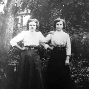 dwie młode dziewczyny w bluzkach i spódnicach, z upiętymi włosami, pozują do fotografii na tle drzew i krzewów