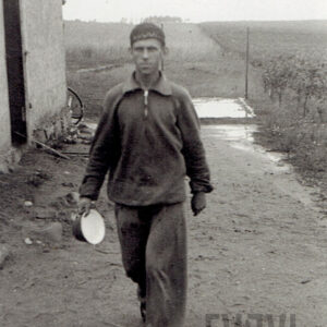 mężczyzna w ubraniu roboczym idzie po piaszczystym terenie, w tle widać pola i fragment murowanego budynku