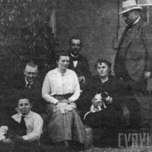 grupa osób - pięć osób siedzi, w tym dwie kobiety, jedna trzyma na kolanach małego pieska, jeden mężczyzna w kapeluszu stoi - na tle ściany budynku z fragmentem okna