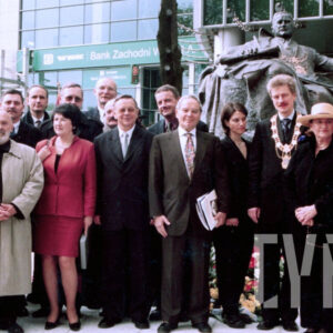 duża grupa ludzi, kobiet i mężczyzn, stoi na tle całopastaciowego pomnika siedzącego mężczyzny, w głębi fragment szklanych elewacji biurowców