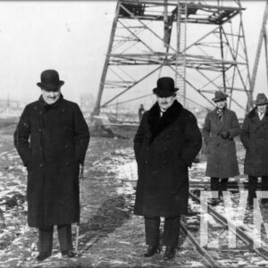 siedmiu mężczyzn w grubych płaszczach i kapeluszach stoi na ośnieżonym polu na tle fragmentu metalowej konstrukcji