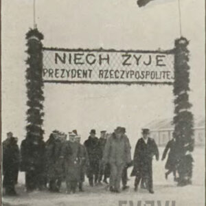 grupa ludzi przechodząca przez prowizoryczną bramę z napisem powitalnym: "Niech żyje prezydent Polski", przyozdobioną kwiatami i flagami