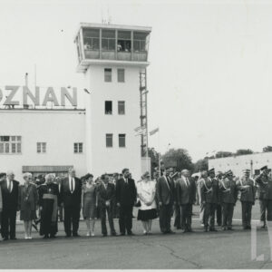 w długim szeregu stoją kobiety i mężczyźni, pięciu mężczyzn w mundurach wojskowych, w głębi budynek z wieżą i napisem Poznań