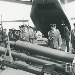 na pierwszym planie wojskowe działo, w głębi mężczyźni w mundurach niosą trumnę, za nimi otwarty właz samolotu