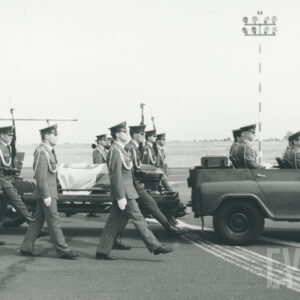 samochód, w którym siedzą żołnierze ciągnie lawetę wojskową na której leży trumna przykryta biało-czerwoną flagą, po obu stronach trumny idą żołnierze 