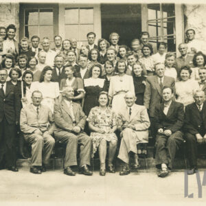 duża grupa kobiet i mężczyzn pozuje przed wejściem do budynku, kobiety maja na sobie letnie sukienki, mężczyźni przeważnie garnitury, za nimi szerokie drzwi wejściowe