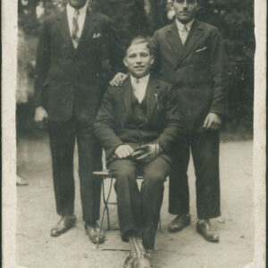 z przodu na krześle siedzi młody mężczyzna, za nim stoi dwóch młodych mężczyzn, cała trójka w garniturach pozuje do zdjęcia na tle drzew, mężczyzna z prawej, trzyma swoją prawą rękę na prawym ramieniu siedzącego mężczyzny