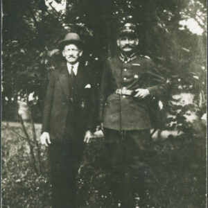 dwóch mężczyzn stoi i pozuje do zdjęcia na tle wysokich krzaków, mężczyzna z lewej w garniturze i kapeluszu, z prawej w mundurze