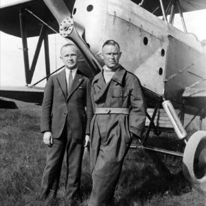 dwóch mężczyzn w średnim wieku, opartych o samolot i pozujących do fotografii, jeden z nich ubranych jest w elegancki garnitur, koszulę i krawat; drugi nosi strój pilota