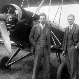 dwóch mężczyzn w średnim wieku, stojących przed samolotem i pozujących do fotografii, ubranych w eleganckie garnitury, koszule i krawaty