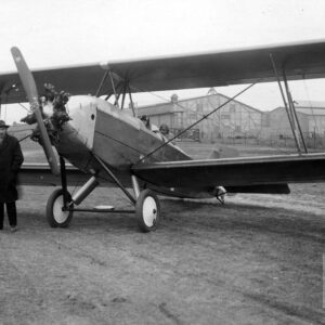 dwóch mężczyzn w płaszczach z kapeluszami na głowach, stojących przy samolocie, w środku którego widać dwóch pilotów, z tyłu budynki przemysłowe