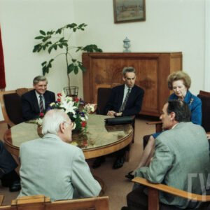 starsza kobieta i grupa starszych mężczyzn w eleganckich ubraniach, siedzący przy okrągłym stole, na którym stoi wazon z kwiatami, na ścianie widoczny fragment godła narodowego