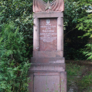 tablica nagrobna o nieregularnym kształcie umieszczona na wysokim schodkowym cokole, z wyrytym cyrylicą napisem w języku rosyjskim, wokół trawa i drzewa