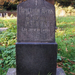 tablica nagrobna z czarnego granitu o nieregularnym kształcie umieszczona na cokole, z wyrytym cyrylicą napisem w języku rosyjskim, wokół trawa i drzewa