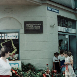 wieńce kwiatów pod tablicą pamiątkową zamontowaną na ścianie budynku z cegły przy skrzyżowaniu ulic