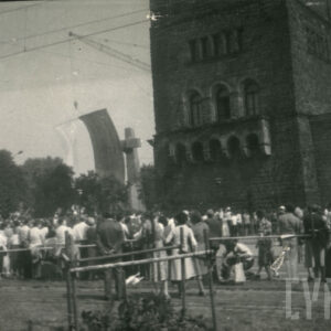 grupa osób stojących tyłem, przyglądających się odsłonięciu pomnika w formie dwóch stalowych krzyży, widoczny też monumentalny budynek z kamienia