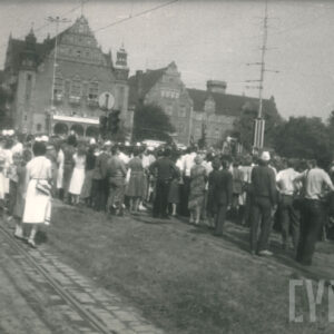 grupa osób stojących tyłem, przyglądających się odsłonięciu pomnika, widoczny też monumentalny budynek z cegieł