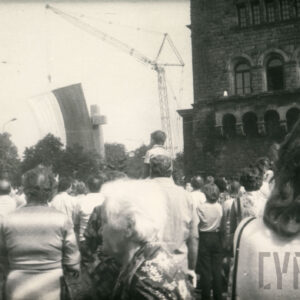 grupa osób stojących tyłem, przyglądających się odsłonięciu pomnika w formie dwóch stalowych krzyży, widoczny też monumentalny budynek z kamienia