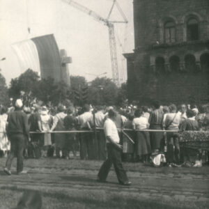 grupa osób stojących tyłem, przyglądających się odsłonięciu pomnika w formie dwóch stalowych krzyży, widoczny też monumentalny budynek z kamienia