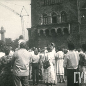 grupa osób stojących tyłem, przyglądających się odsłonięciu pomnika w formie dwóch stalowych krzyży, widoczny też monumentalny budynek z kamienia
