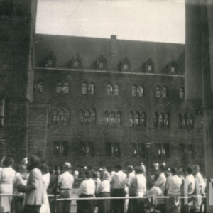 grupa osób stojących tyłem, przyglądających się odsłonięciu pomnika, widoczny też monumentalny budynek z kamienia